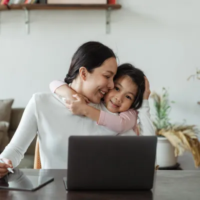 Young girl hugging woman whilst at a table on a laptop