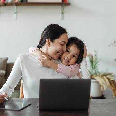 Young girl hugging woman whilst at a table on a laptop